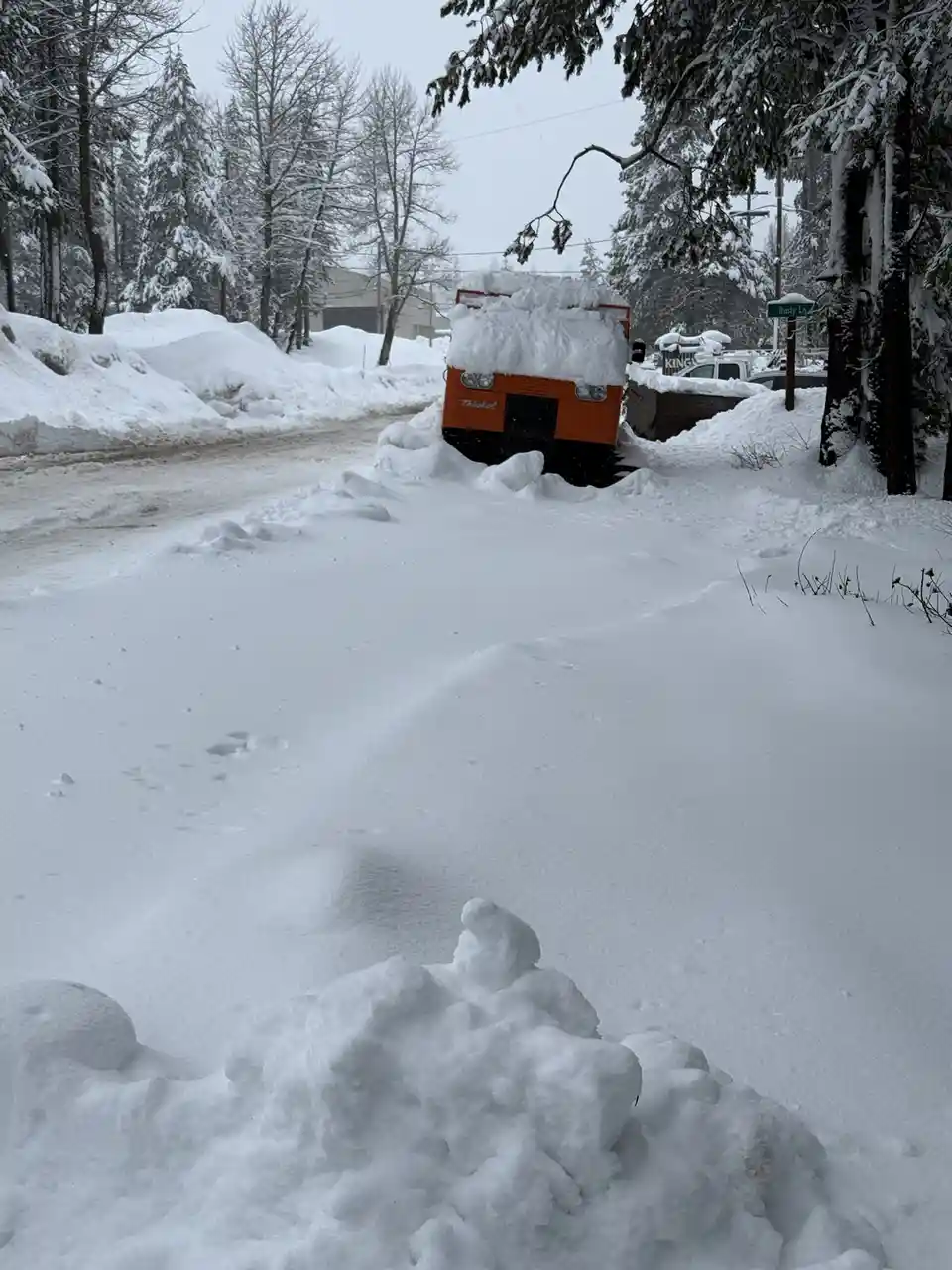 A snow-covered orange snowplow truck is buried in deep snow on a snowy street, with snowdrifts piled high around it and a…