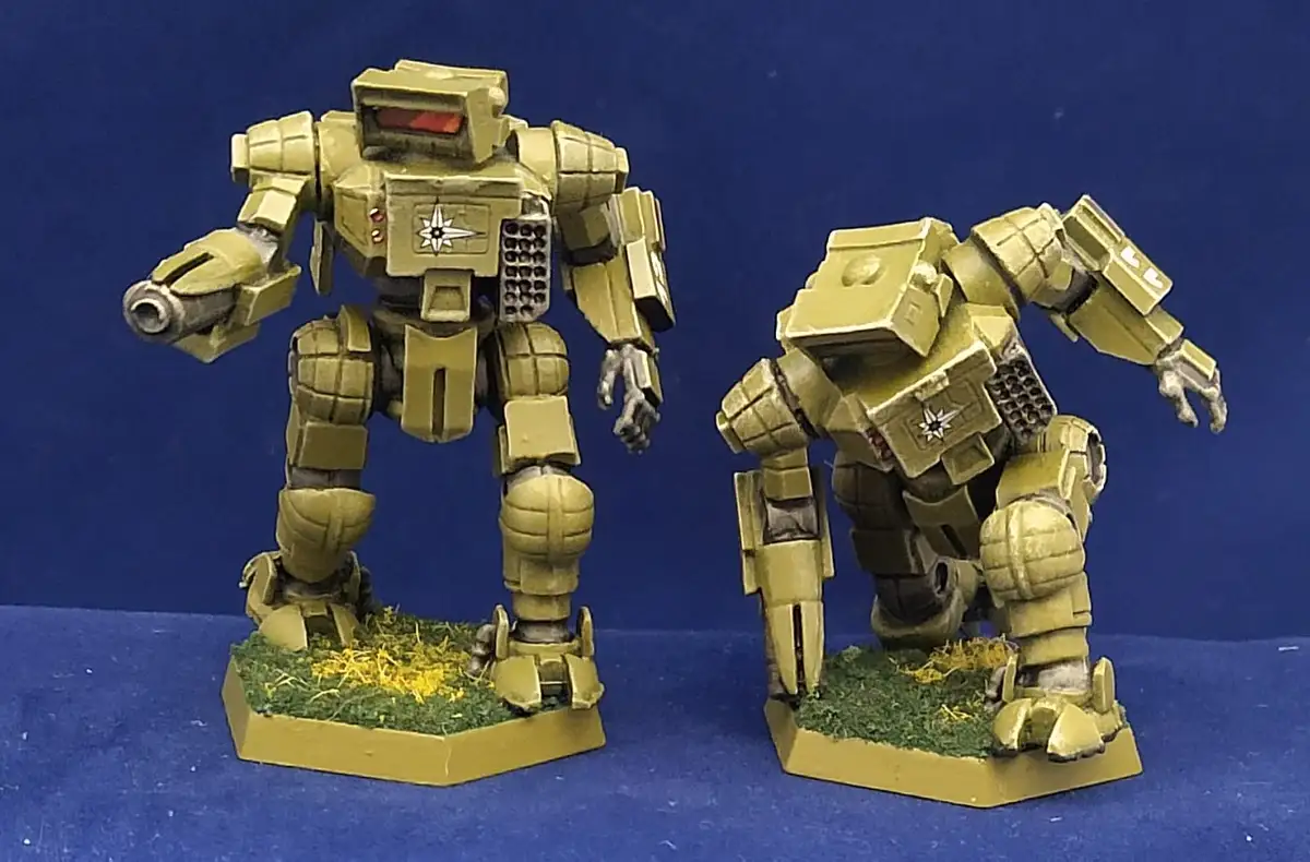 Two painted yellow humanoid robot miniatures stand on grassy bases against a blue background. The robot on the left stands…