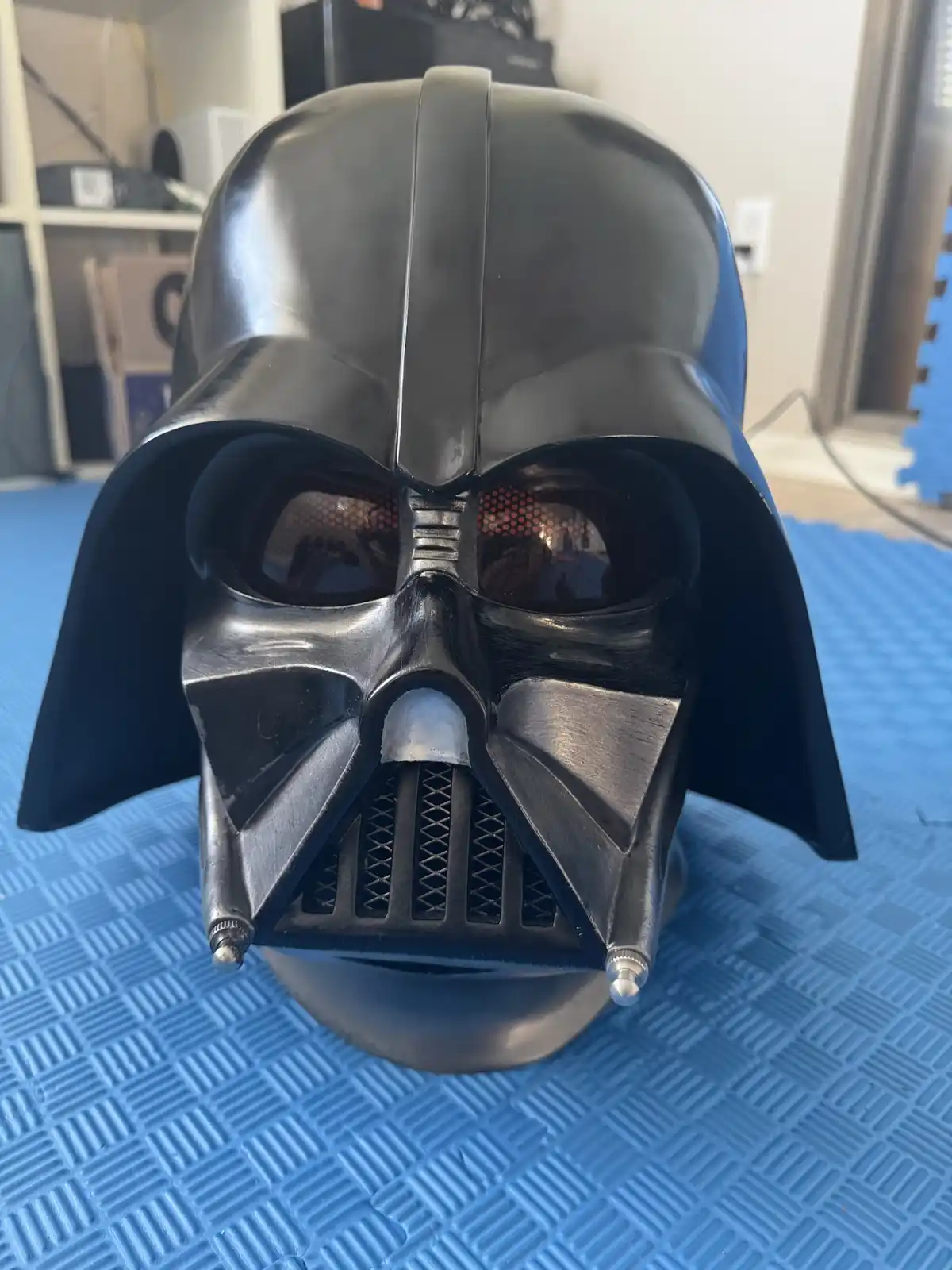 A close-up photo shows a black Darth Vader helmet resting on a blue textured mat, with a background of shelves and a…