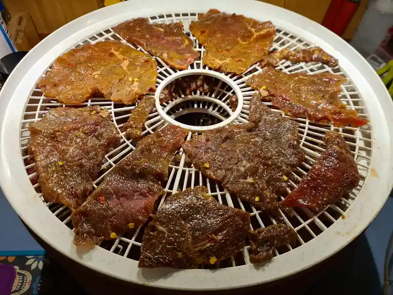 A white circular food dehydrator tray is filled with numerous raw, marinated meat slices, seasoned with visible red pepper…