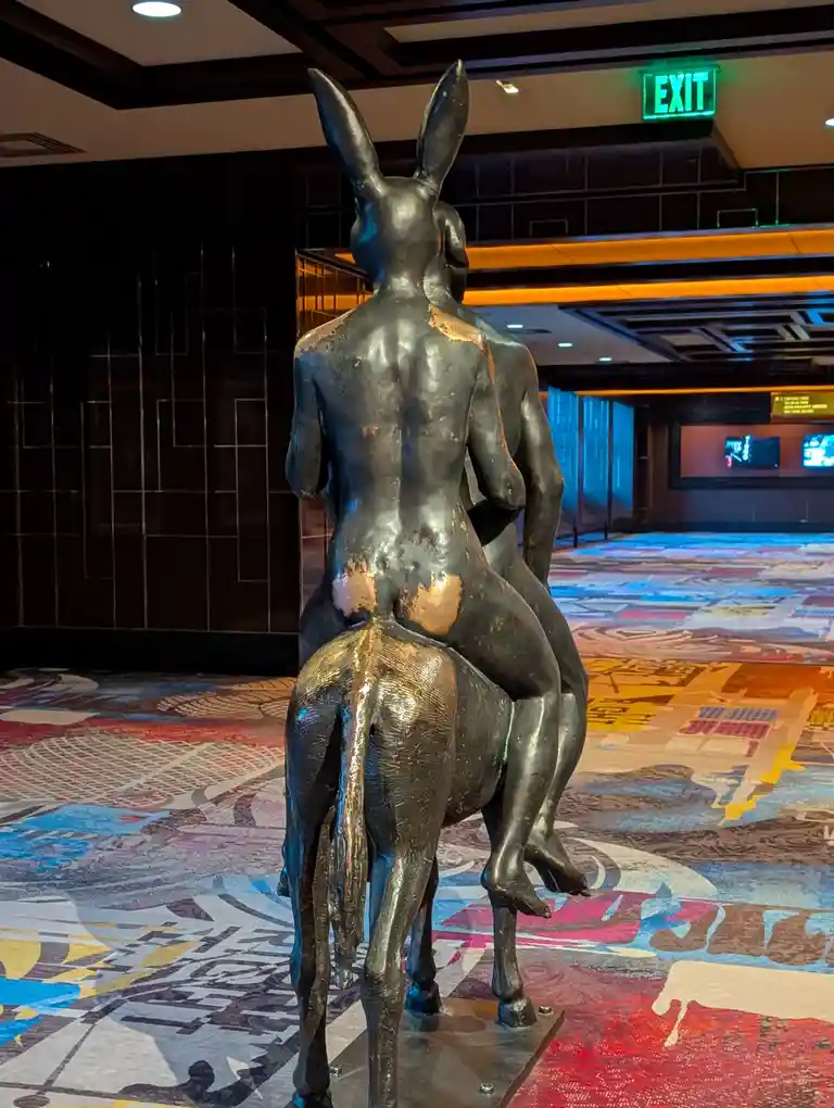 A bronze statue of a nude human figure with rabbit ears riding a horse, viewed from behind, stands in a hotel lobby with a…