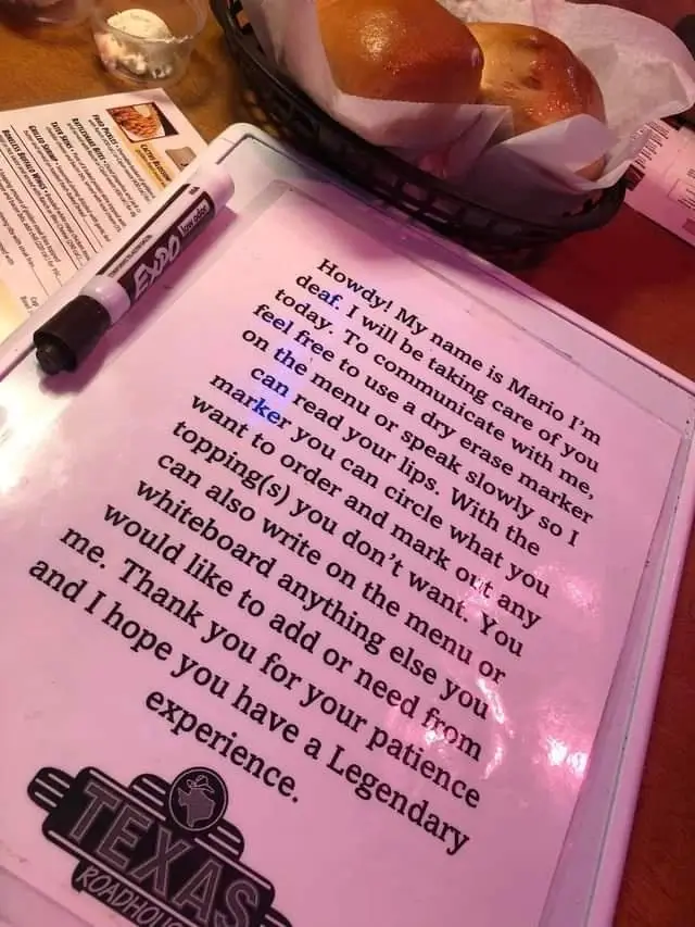 A pink dry-erase board at a Texas Roadhouse restaurant displays a handwritten note from a deaf server named Mario…