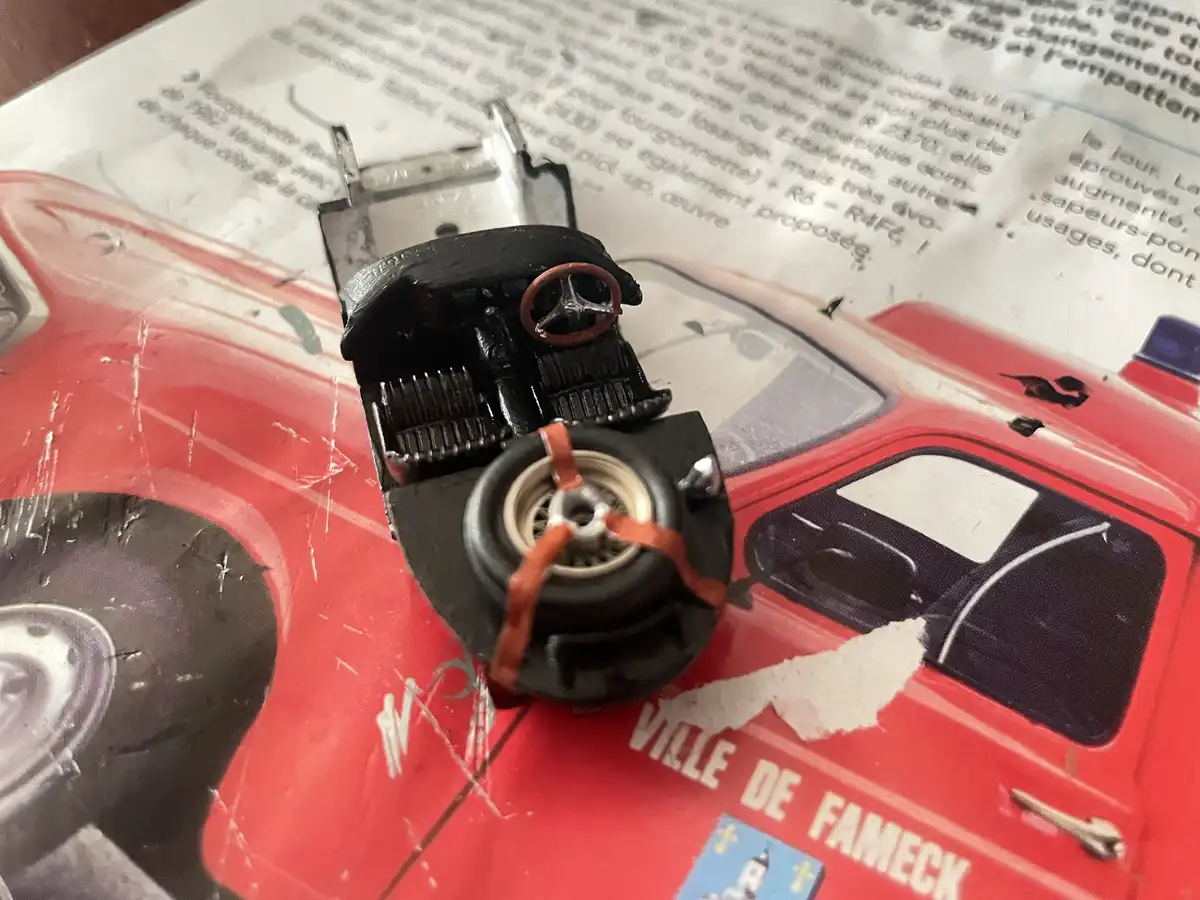 A small, crudely assembled toy car made from a toaster, with a wheel strapped on with orange bands, sits atop a torn red…