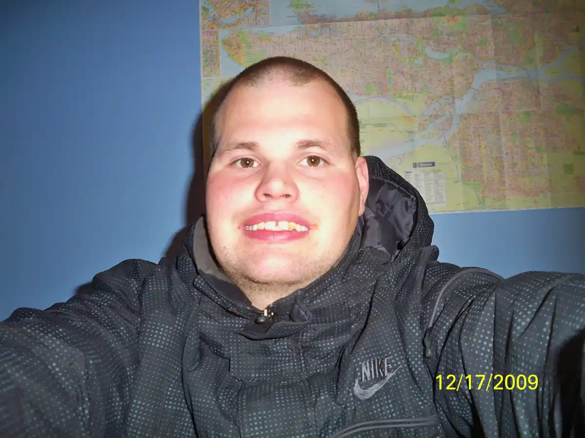 A man in a dark polka-dotted Nike jacket takes a selfie with a wide, toothy grin, standing in front of a large Vancouver…