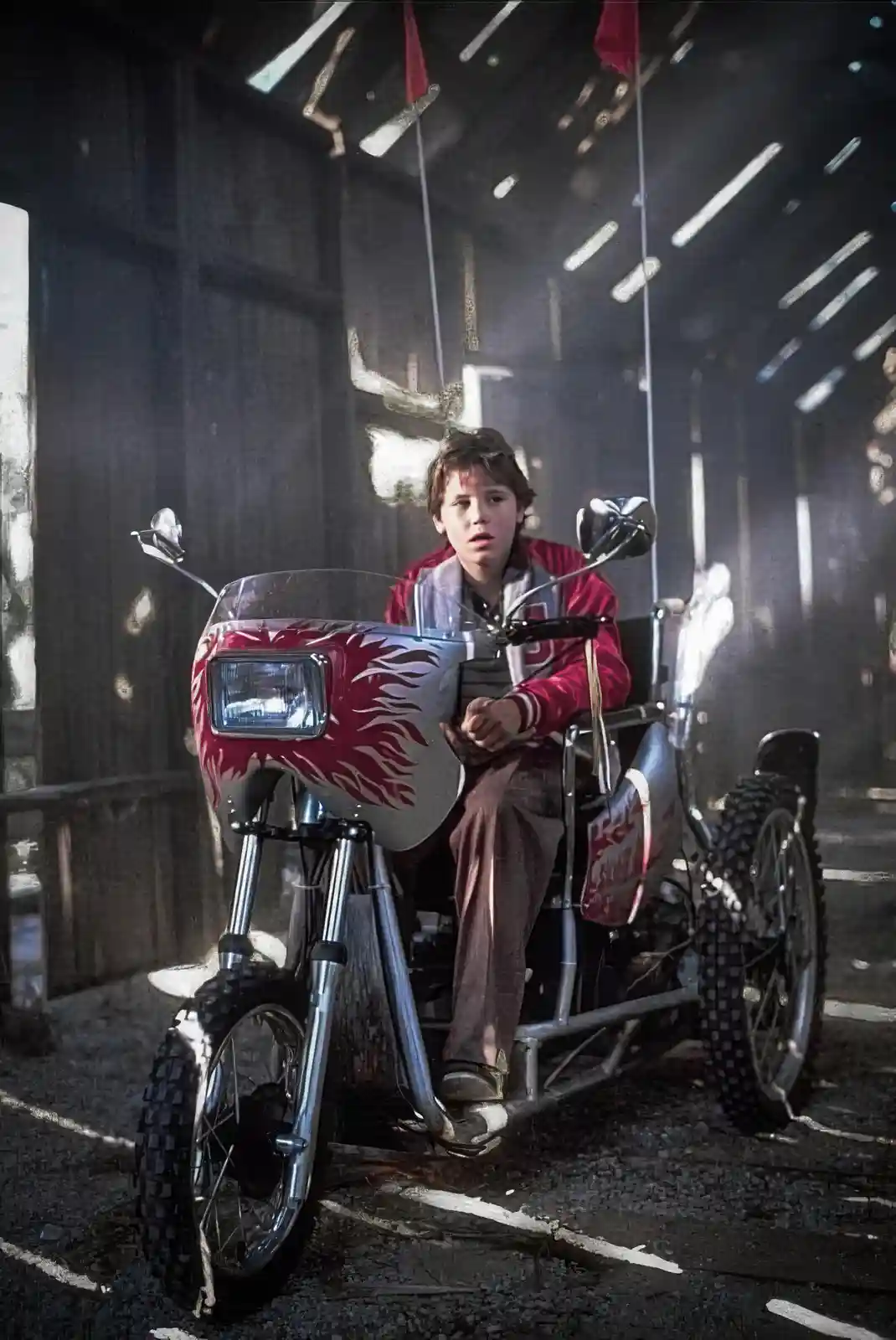 A young boy with a surprised expression sits on a custom-built, flame-decorated three-wheeled motorcycle inside a dimly lit…
