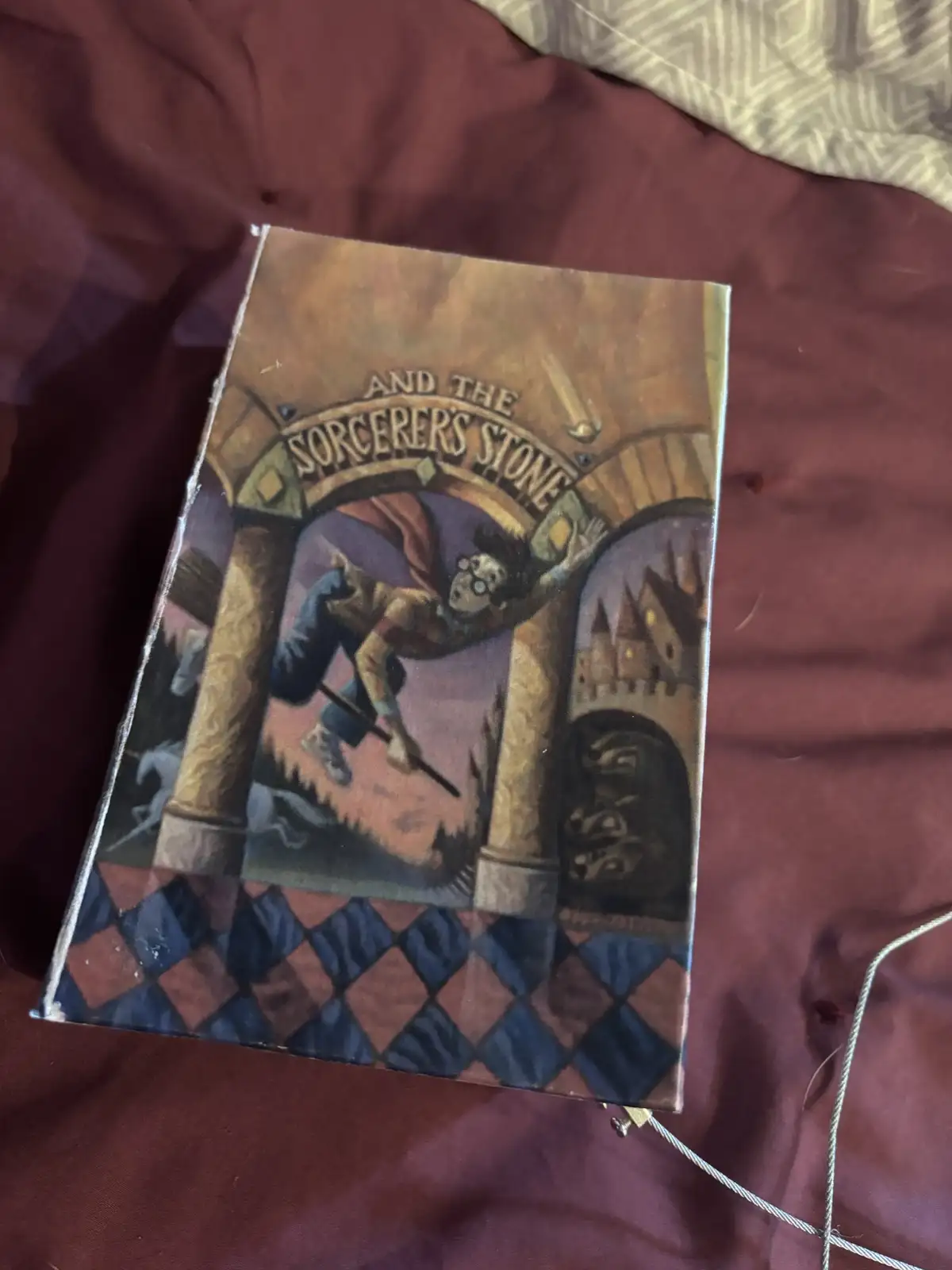 A physical copy of the book 'Harry Potter and the Sorcerer's Stone' is shown lying on a maroon fabric surface, with its…