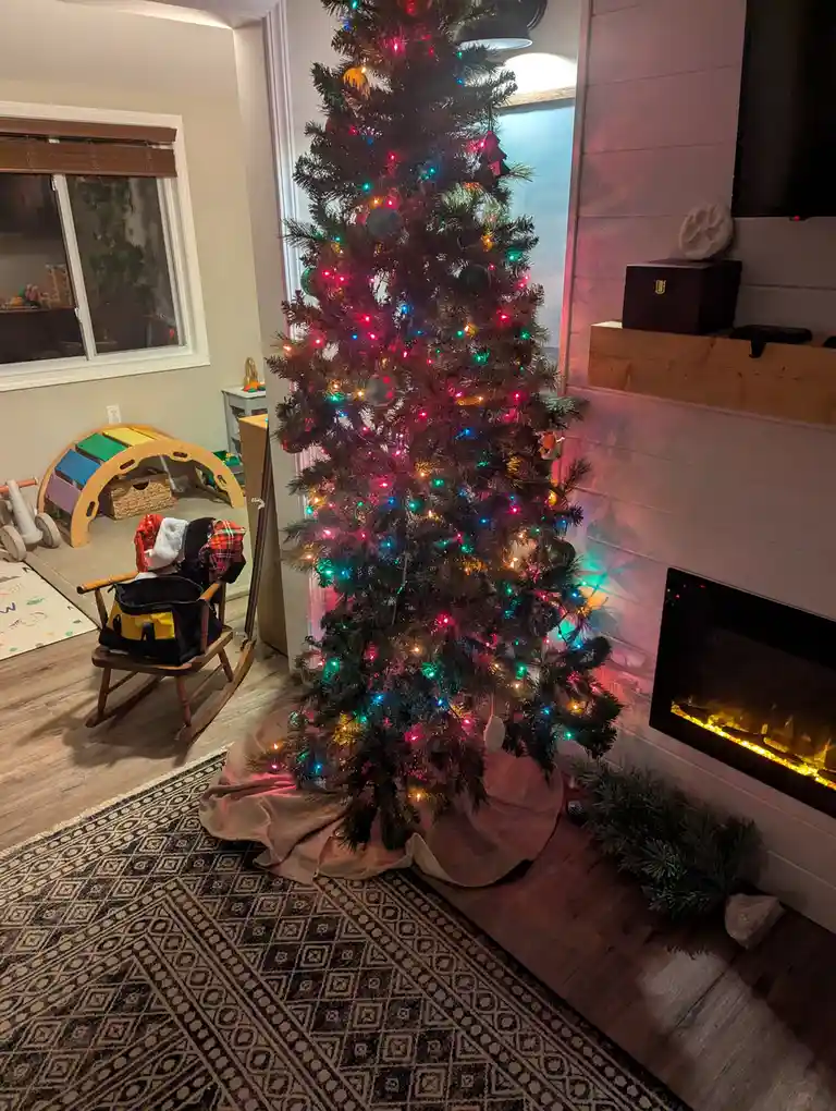 A decorated Christmas tree with colorful lights and ornaments stands in a living room next to a fireplace. In the…
