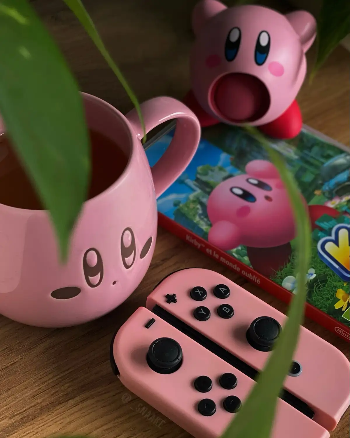 A cozy setup featuring pink Kirby-themed items: a mug with Kirby's face, a Kirby figurine, a pink Nintendo Switch Joy-Con…
