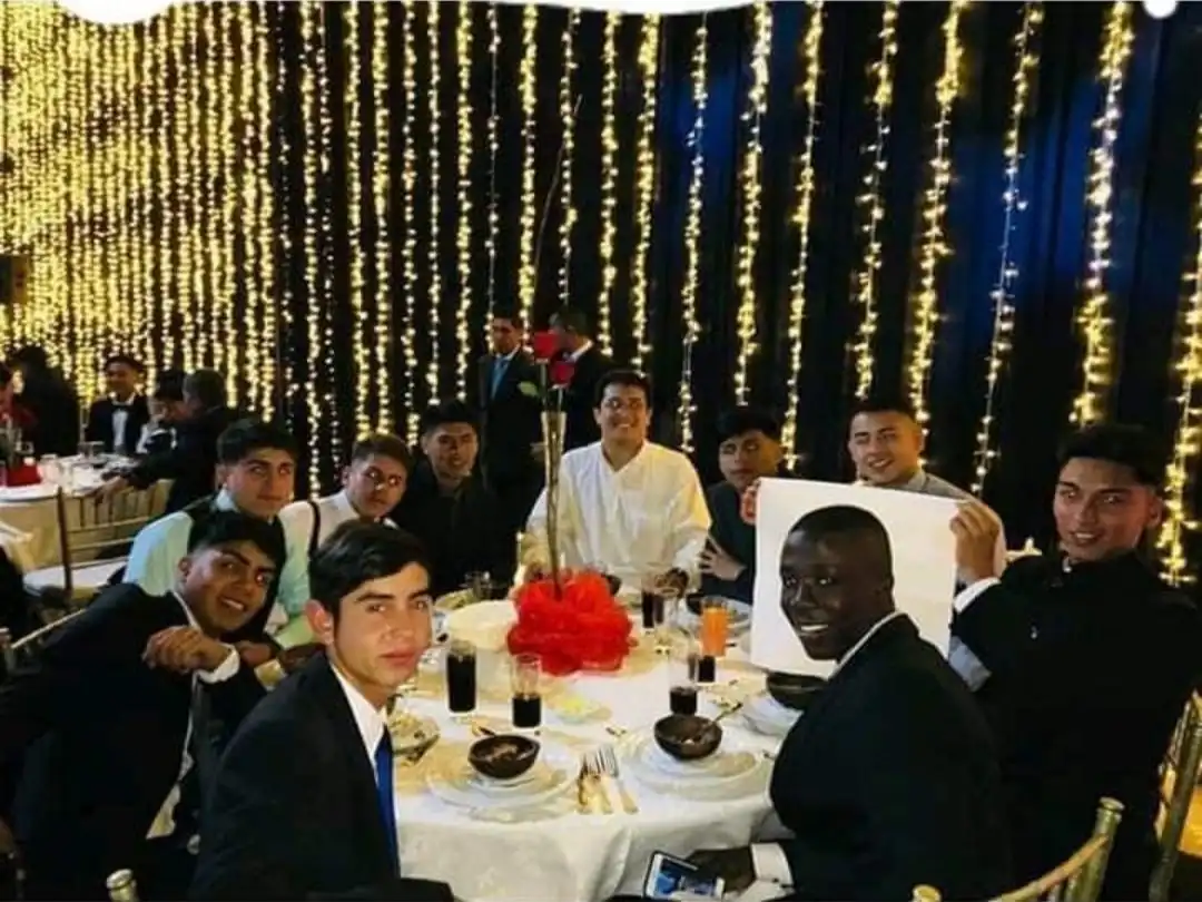 A group of young men in formal wear are seated at a banquet table with string lights in the background. One man holds up a…