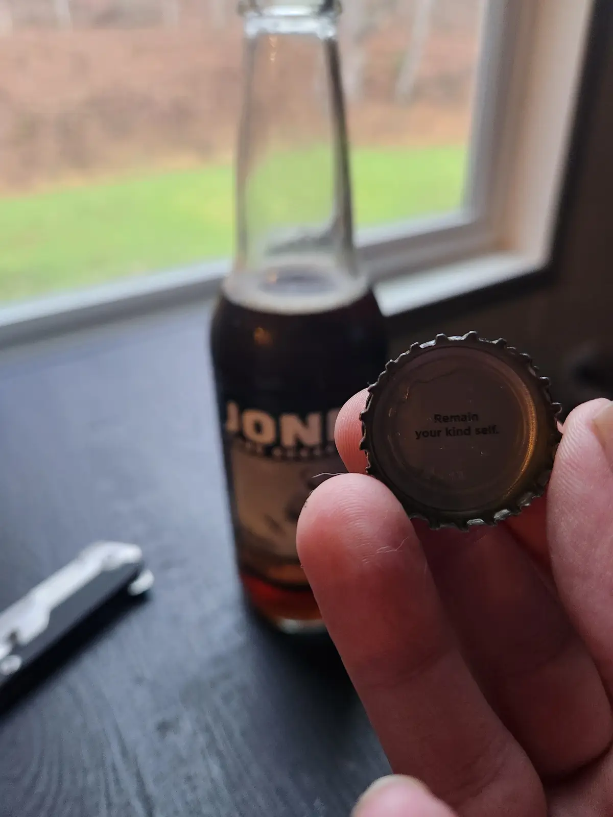 A hand holds a metal bottle cap with the text 'Remain your kind self.' printed on it, in front of a dark beer bottle labeled…