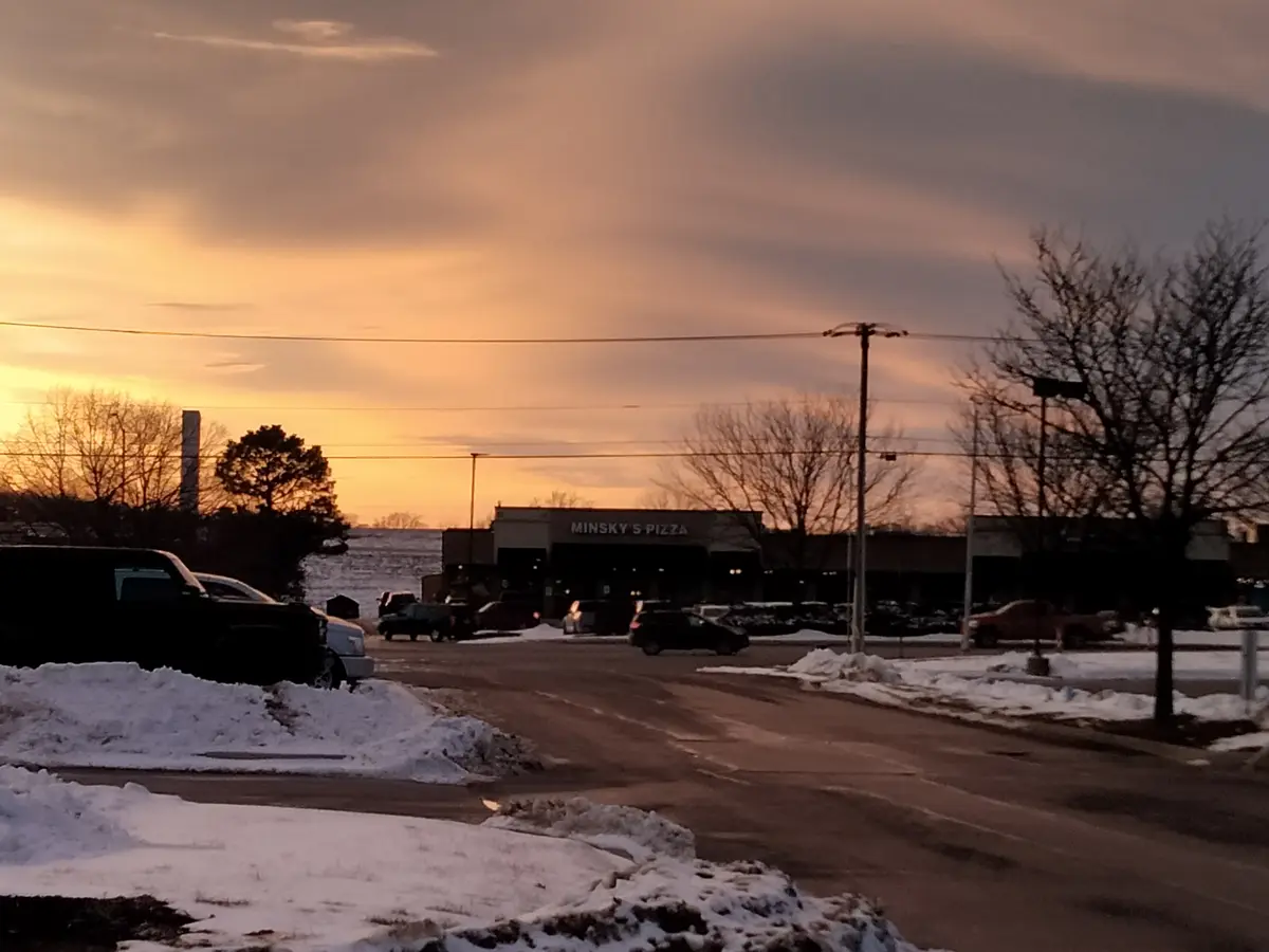A photo of a snowy parking lot at sunset with a building labeled 'MINSKY'S PIZZA' in the background, cars parked, and bare…