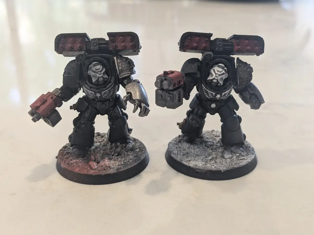 Two heavily armored, custom-painted miniature figures resembling Space Marines stand side-by-side on textured bases. Both…