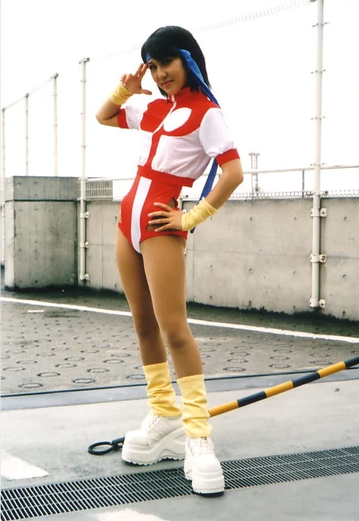 A person in a red and white superhero-style costume with yellow gloves and boots poses on a rooftop, making a peace sign…