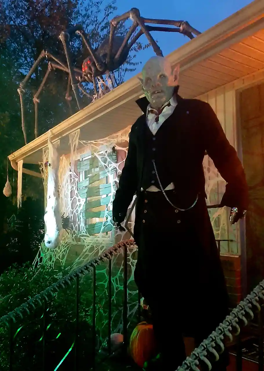 A Halloween display features a large, pale, bald, elf-like figure in a black suit standing on a porch, with a giant skeletal…