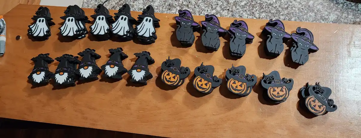 Several stacks of Halloween-themed 3D-printed charms are arranged in four rows on a wooden surface. The top row features…