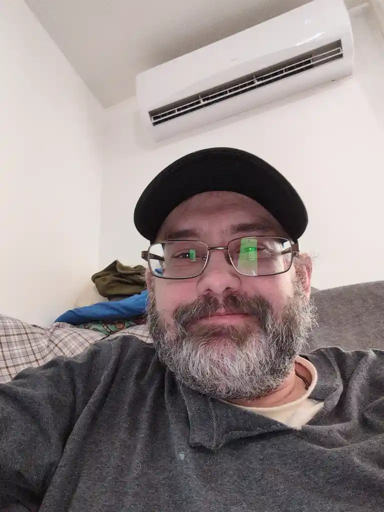 A man with a gray beard, glasses, and a black cap takes a selfie while sitting on a couch, with a white wall-mounted air…
