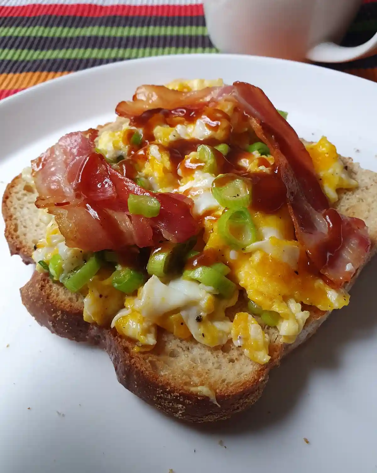 A close-up photo of a slice of toasted bread topped with scrambled eggs, crispy bacon strips, chopped green onions, and a…