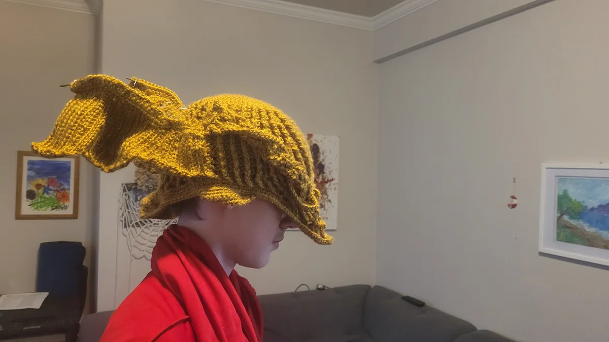 A person in a red robe is shown in profile, wearing a large, yellow, knitted hat that is visibly unfinished and has knitting…