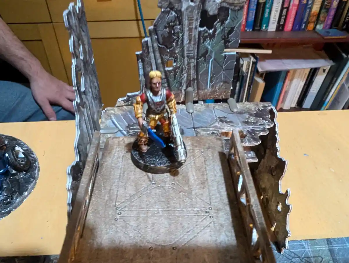 A painted miniature fantasy warrior figure stands on a detailed, ruined castle-themed gaming terrain with stone walls and a…