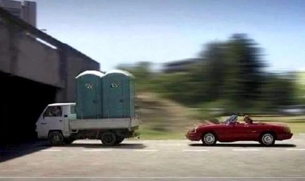 A white truck carrying two green portable toilets drives past a red convertible with two people inside. The background is…