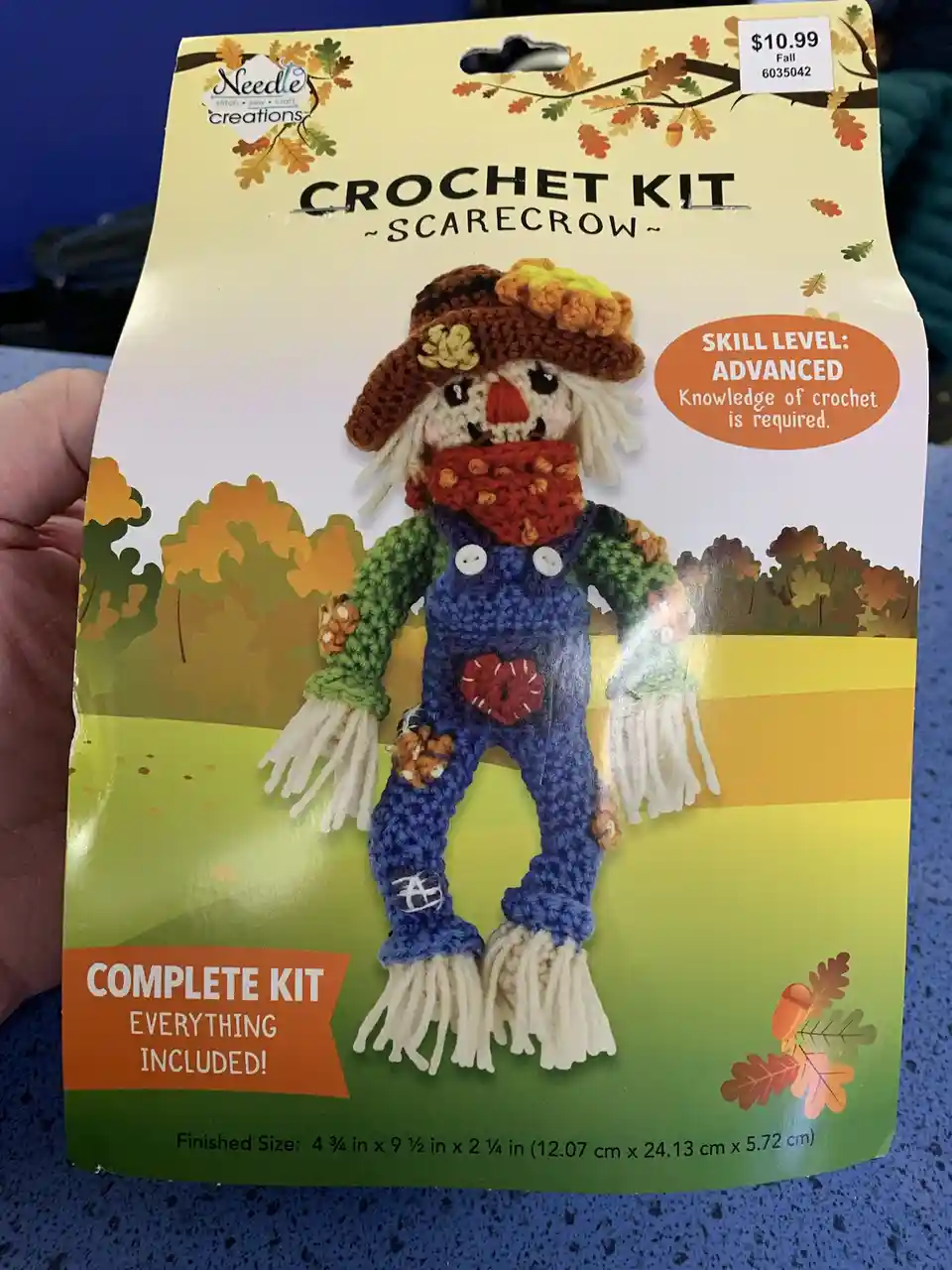 A hand holds a 'Needle Creations' crochet kit package for a scarecrow, which features a small, crocheted scarecrow wearing a…