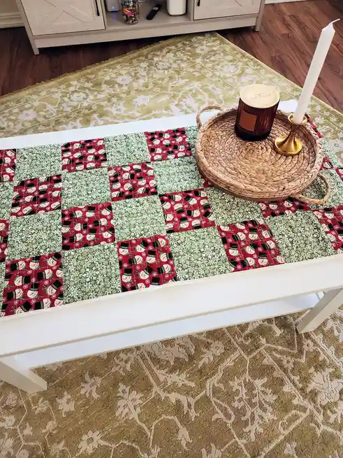 A white coffee table is covered with a handmade patchwork quilt featuring alternating red and green squares with snowman…