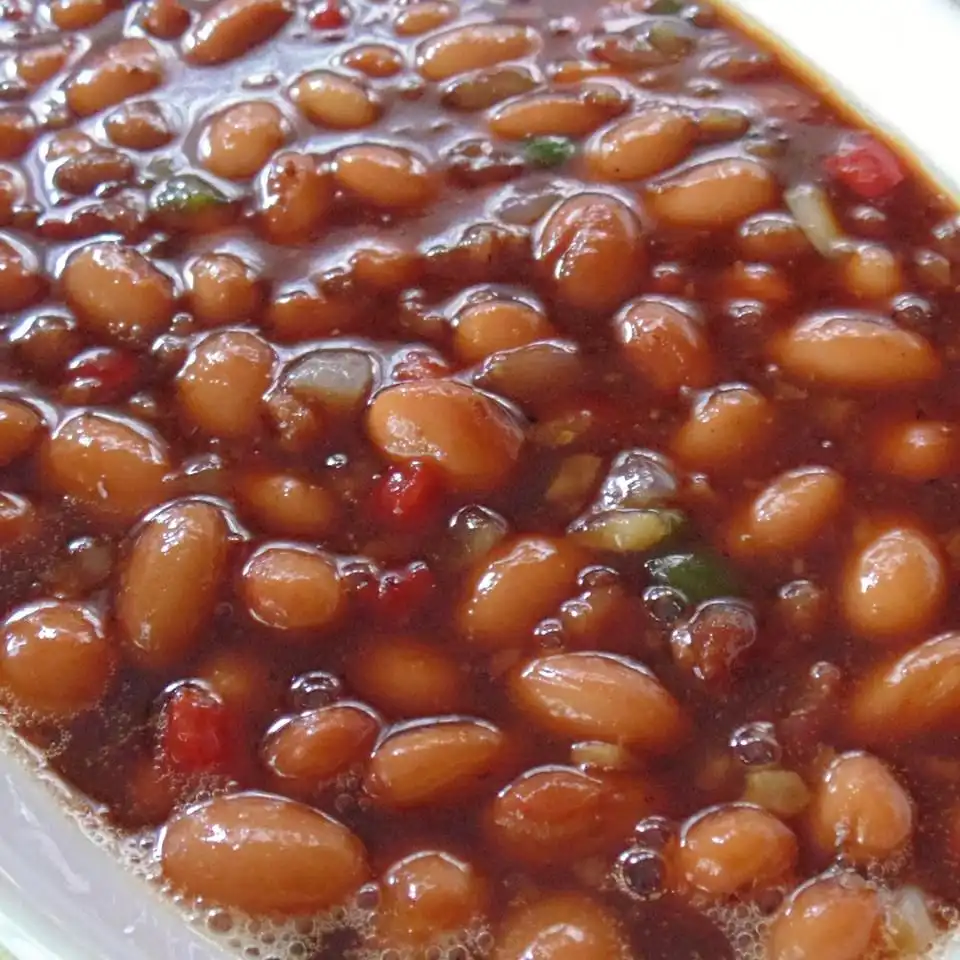 A close-up photo of a bowl of baked beans in a dark, glossy sauce with visible chunks of red and green peppers and onions…