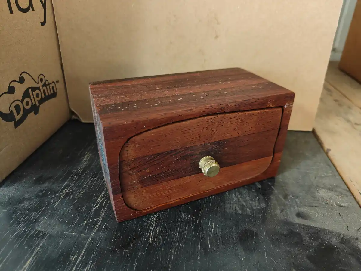 A small, dark wood box with a brass knob sits on a scratched black surface, next to a cardboard box with a 'Dolphin' logo…