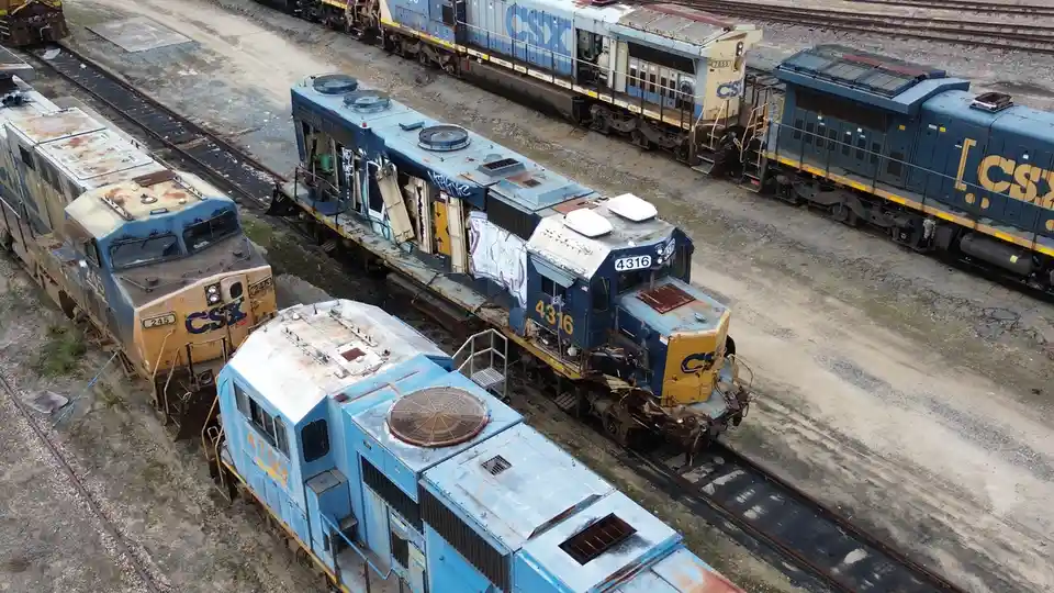 An aerial view shows several rusty, abandoned CSX locomotives sitting on railway tracks in a yard. One train car, number…