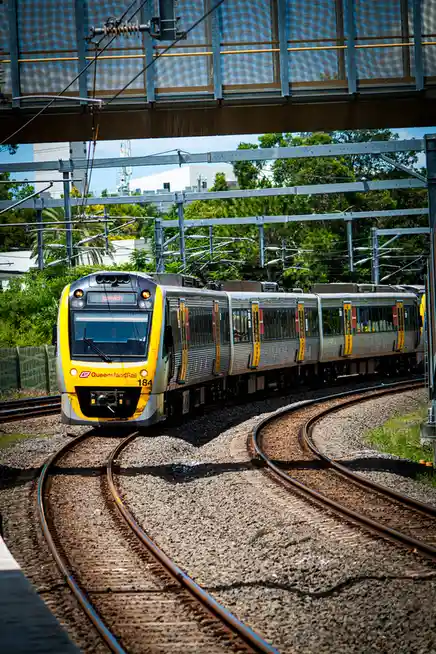 A yellow and silver Queensland Rail train, numbered 184 and headed to Ipswich, is shown on curved tracks under an overhead…