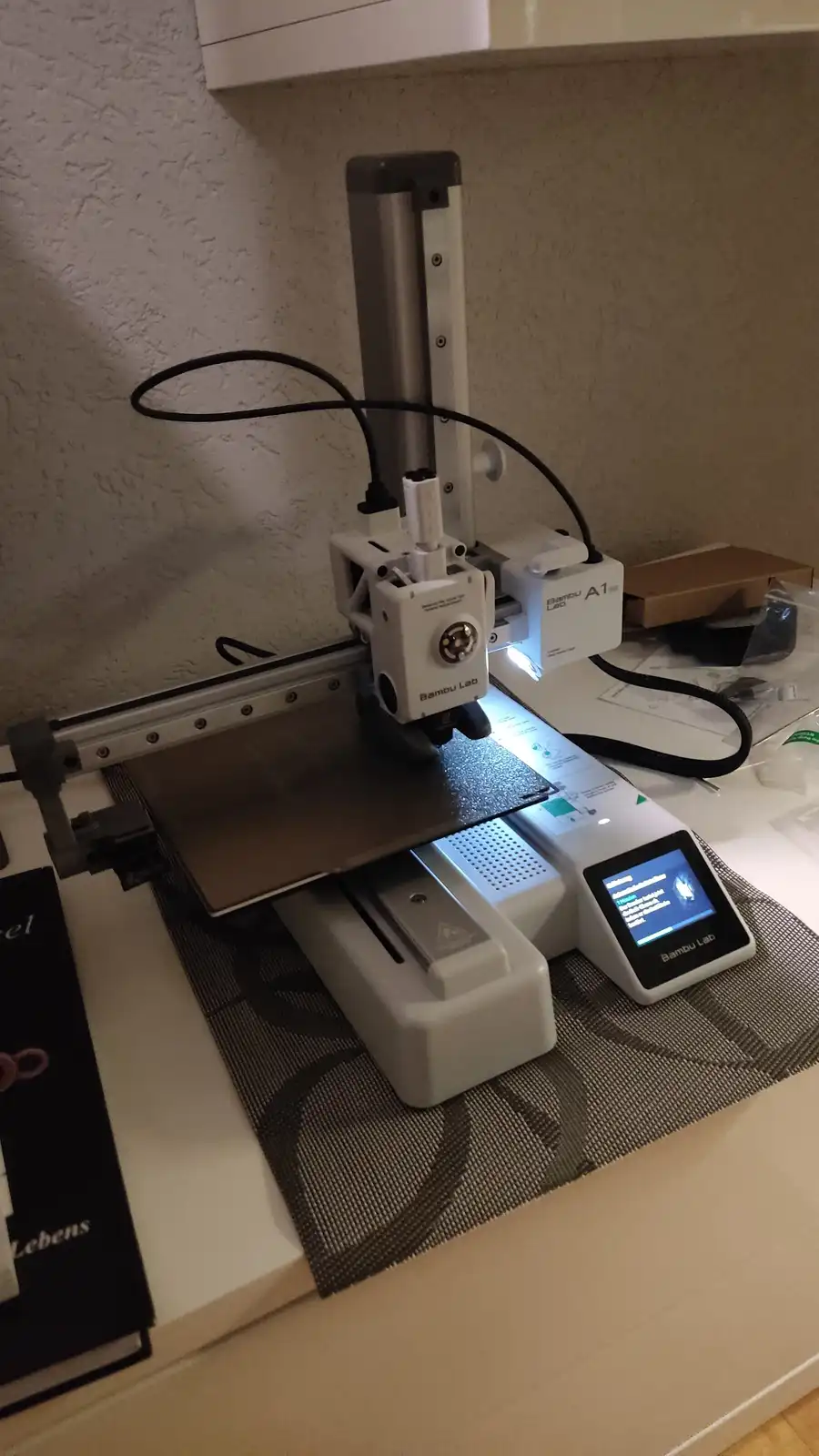 A white Bambu Lab A1 3D printer sits on a kitchen counter, its print head poised over a build plate with a glowing blue…