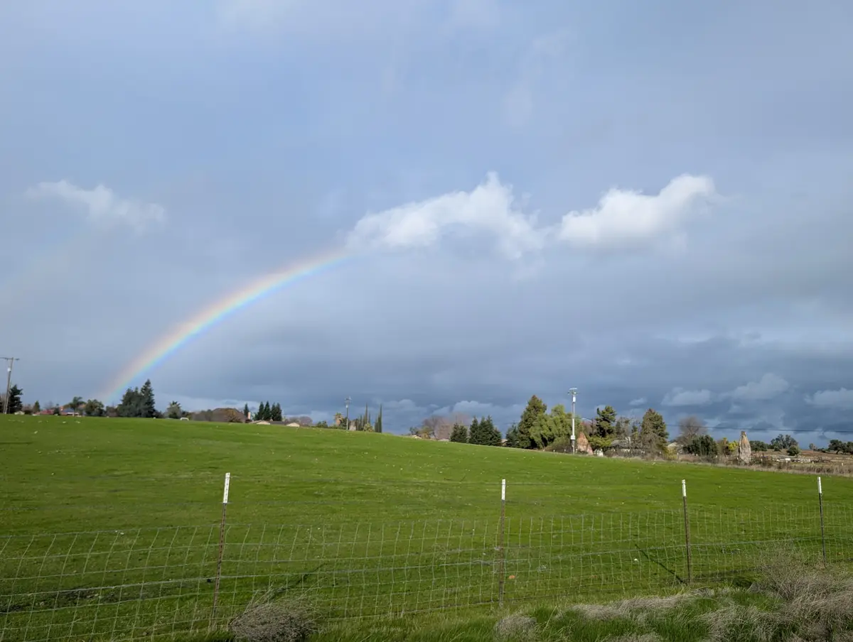 A wide shot of a green grassy field with a wire fence in the foreground, under a partly cloudy sky with a vivid rainbow…