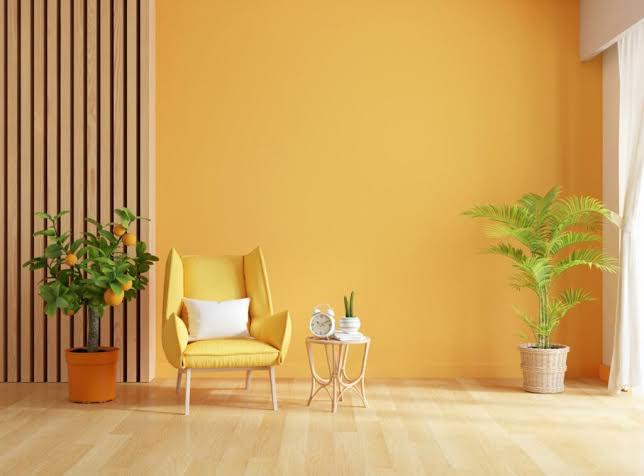 A bright, minimalist room with warm orange walls, a yellow armchair with a white pillow, a small side table with a clock and…