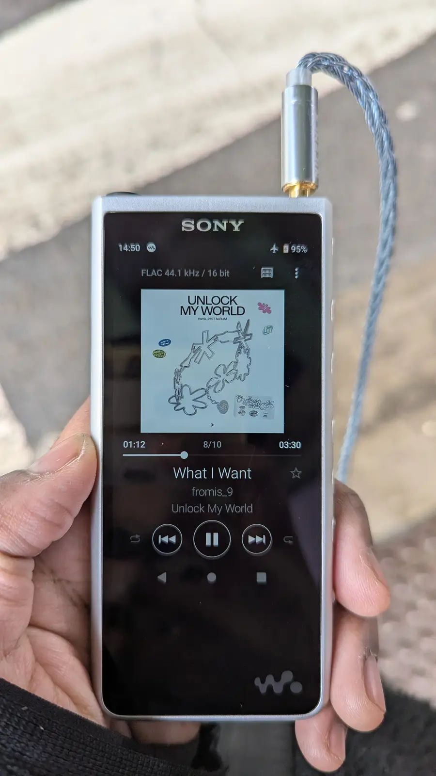 A hand holds a silver Sony Walkman-style portable music player displaying the song 'What I Want' by fromis_9 from the album…