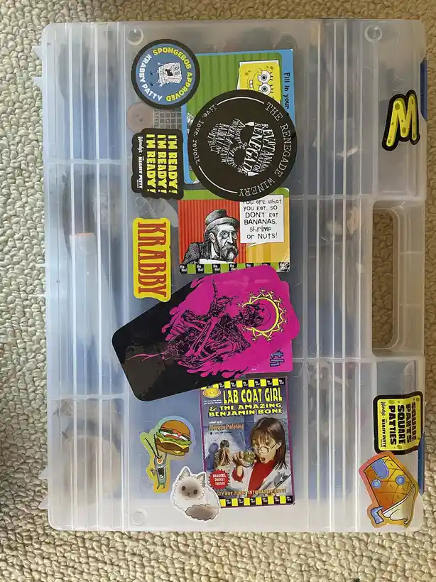 A clear plastic storage box covered in various stickers, including SpongeBob SquarePants-themed ones, a 'Lab Coat Girl' book…