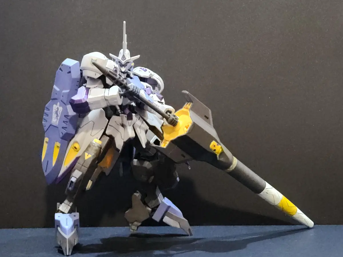A white, purple, and yellow plastic robot model, resembling a Gundam, stands in a dynamic pose holding a large rifle-like…