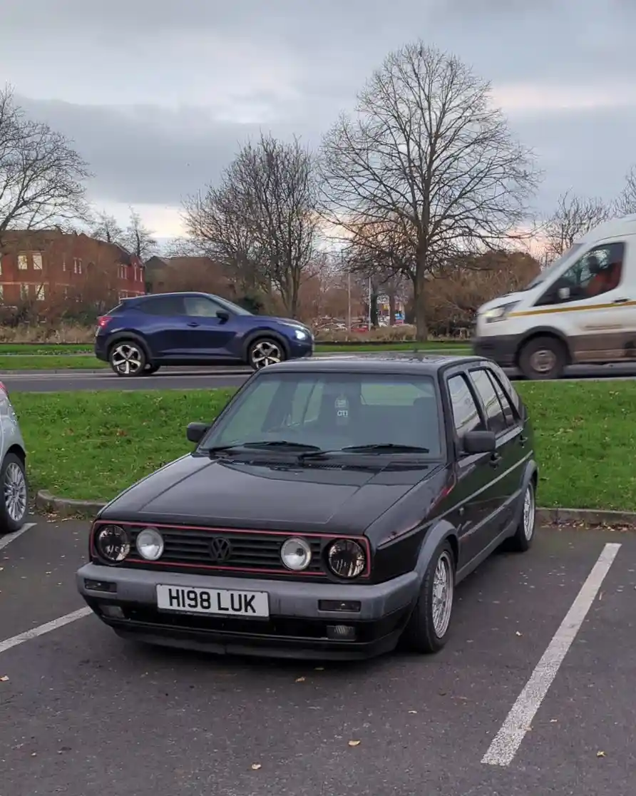 A black 1990s Volkswagen Golf GTI is parked in a lot with license plate 'HI98 LUK', humorously implying the car is 'hi98'…