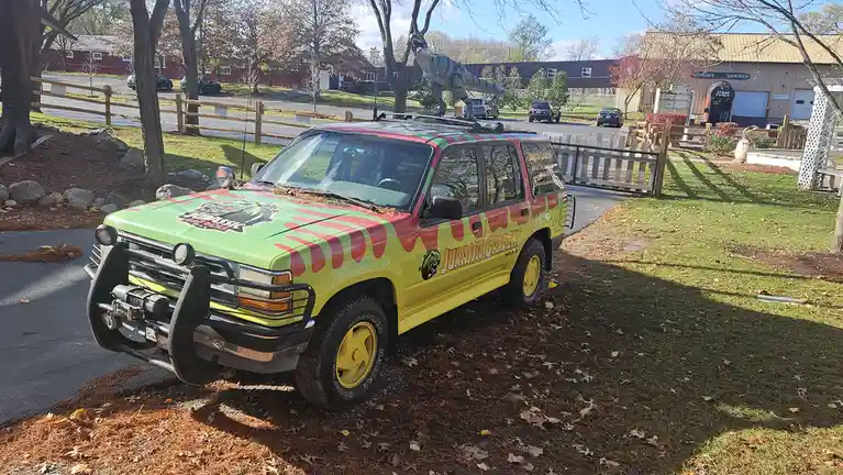 A bright green and yellow SUV with 'Jurassic Gardens' branding and dinosaur-themed decals is parked on a grassy area with…