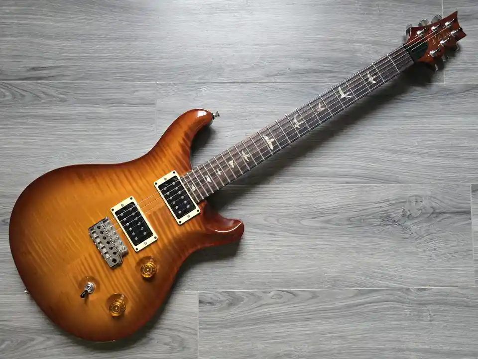 A sunburst-finished electric guitar with bird-shaped inlays on the fretboard lies diagonally on a gray wood-patterned floor…