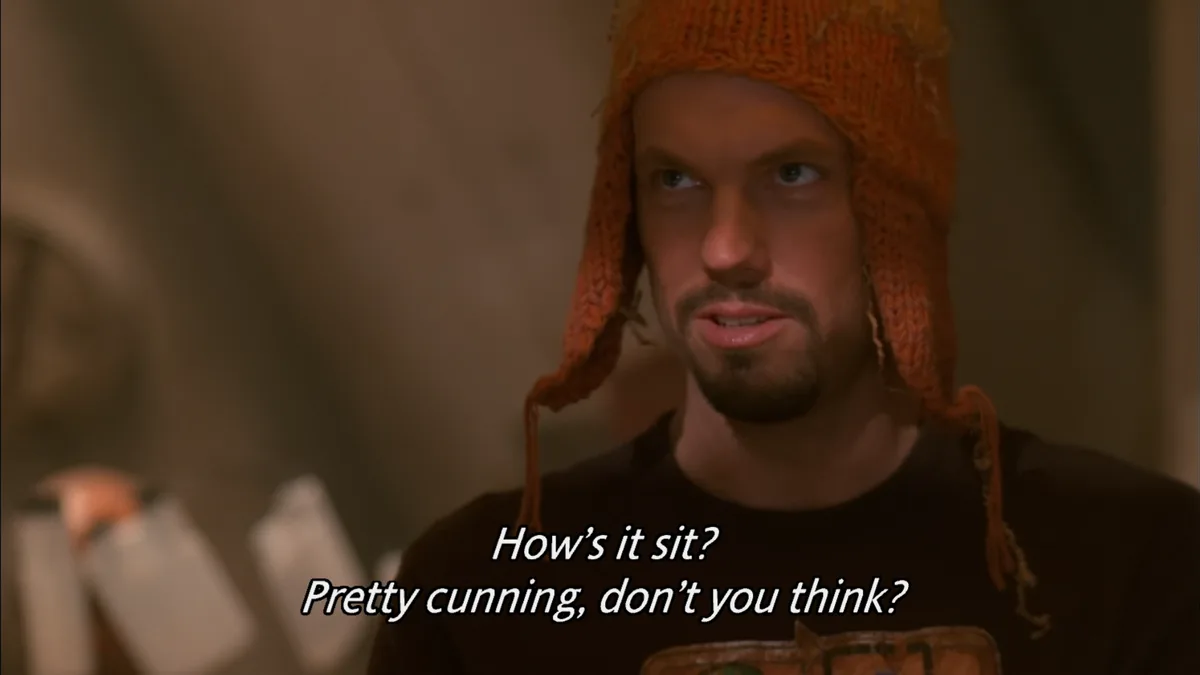 A man wearing a worn orange knitted hat with ear flaps looks sideways with a smug expression. The overlaid text reads 'How's…