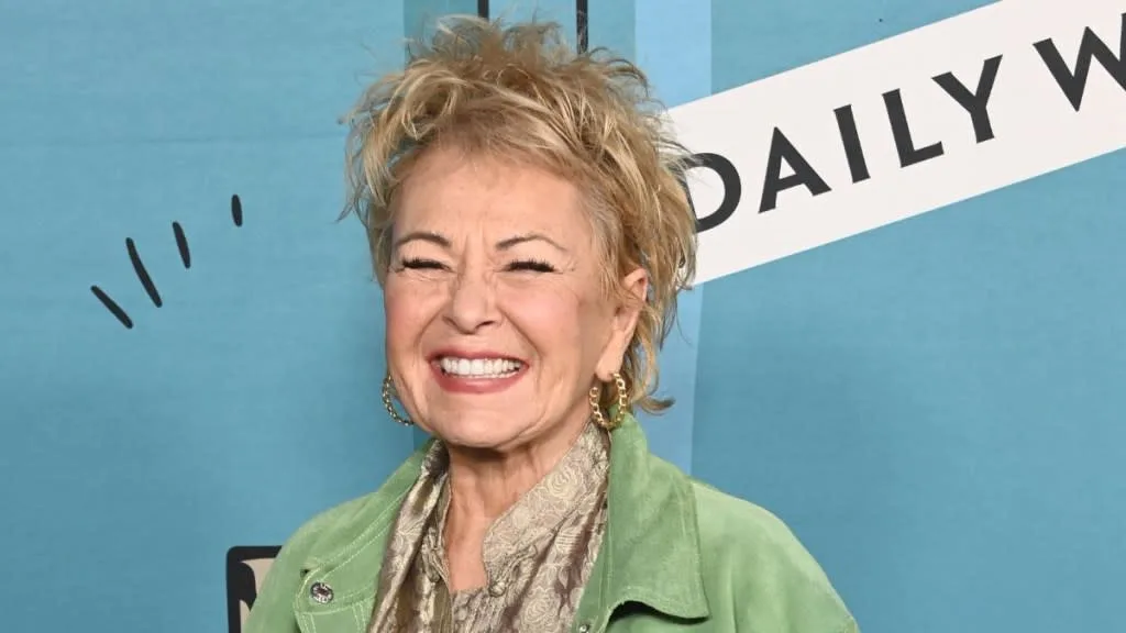 a woman with short blonde hair, wearing a green jacket and gold hoop earrings, is smiling broadly with her eyes squeezed…