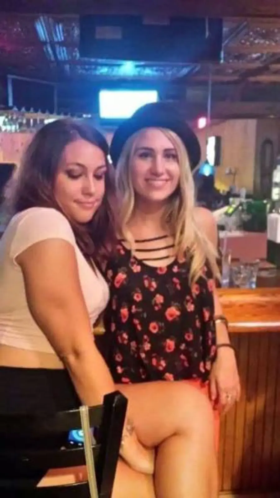 Two young women are posing together in a dimly lit bar setting. The woman on the left, with dark hair, is leaning her head…