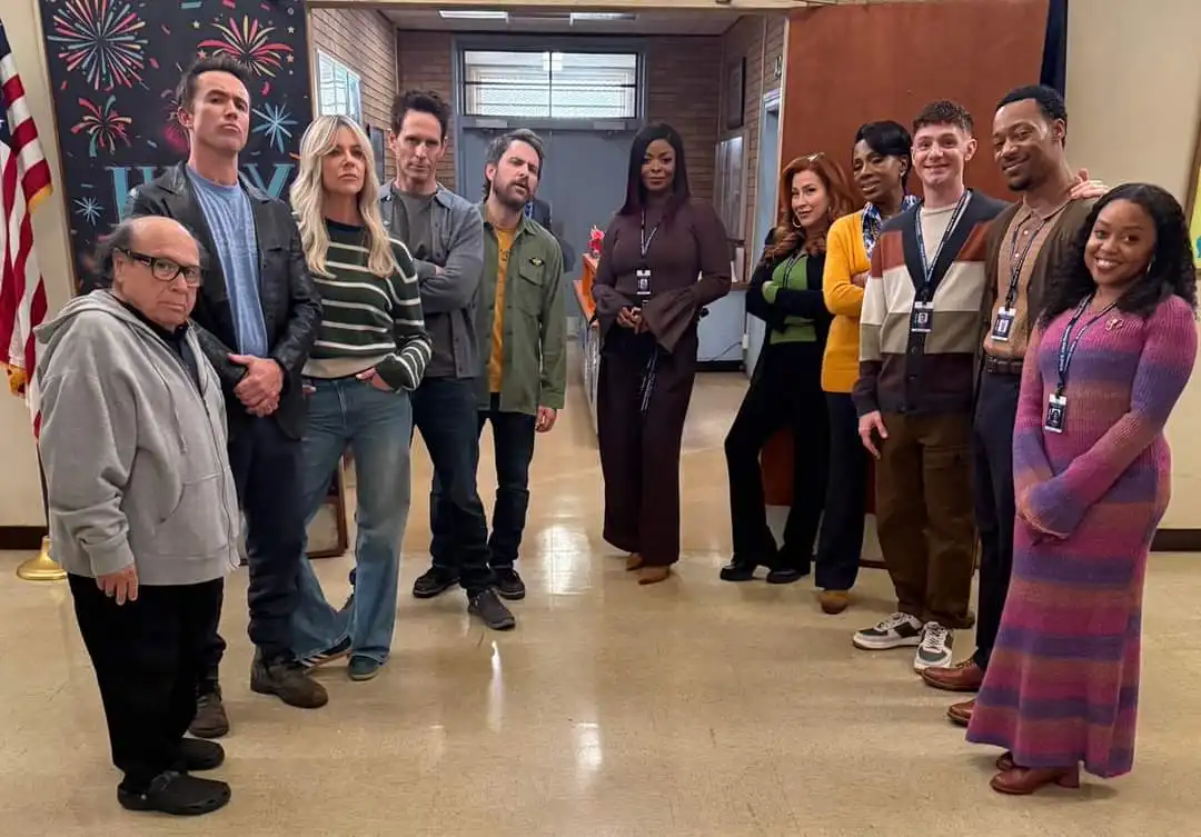 A group of people, including actor Larry David in a grey hoodie and glasses, stand in a school hallway. Most are wearing…