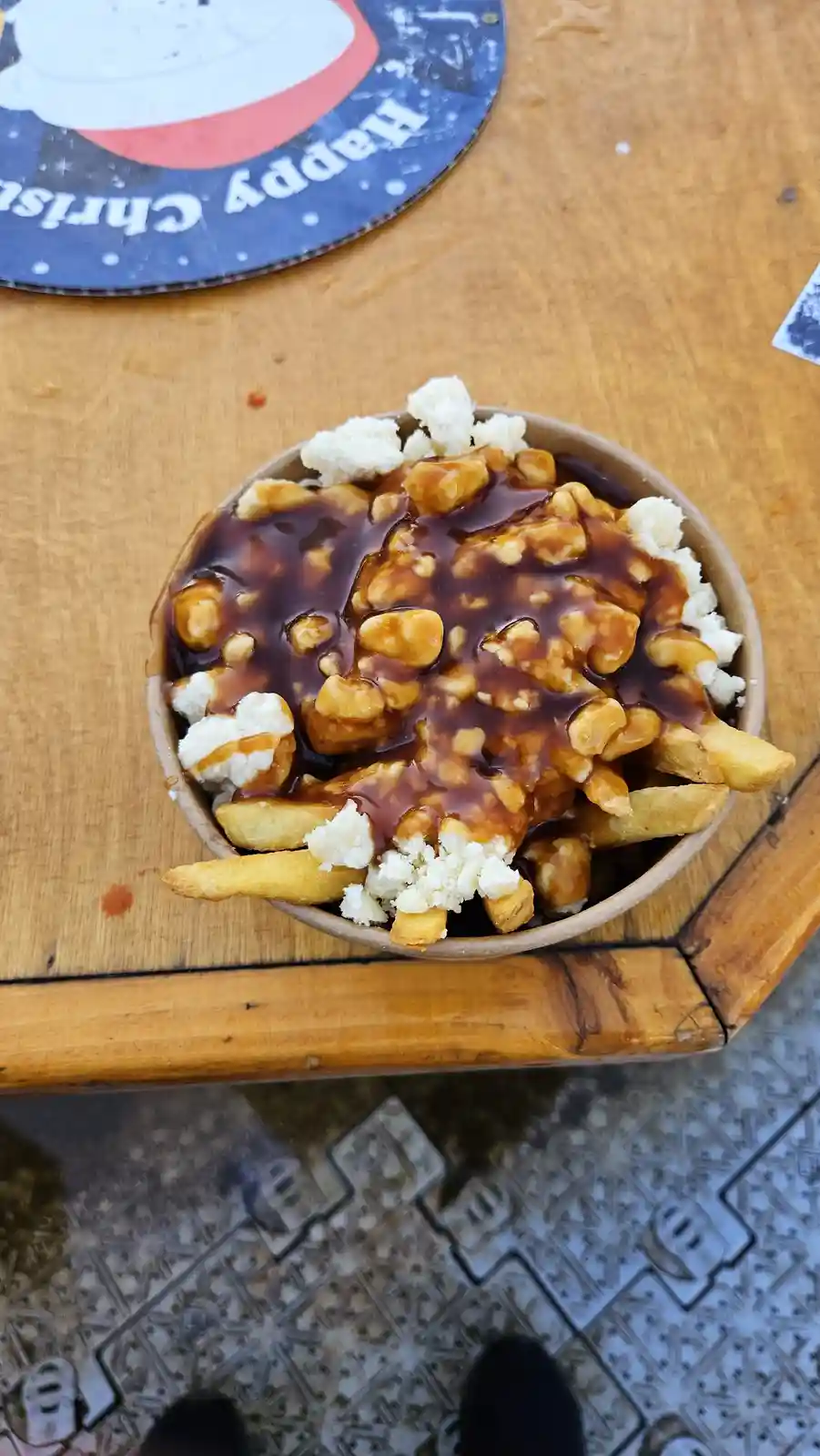 A top-down photo of a paper bowl filled with french fries, white cheese curds, and brown gravy, sitting on a wooden table…