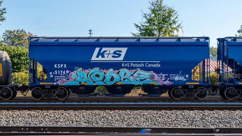 A blue K+S Potash Canada freight train car sits on tracks with trees and a clear sky in the background. The side of the car…