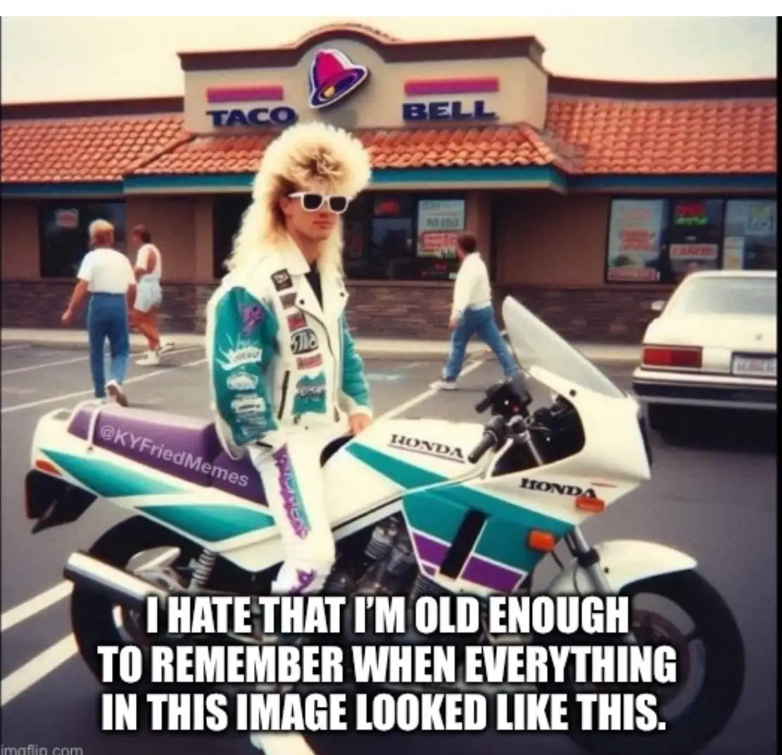 A person with a large, teased blonde mullet and white sunglasses sits on a white and teal Honda motorcycle in front of a…