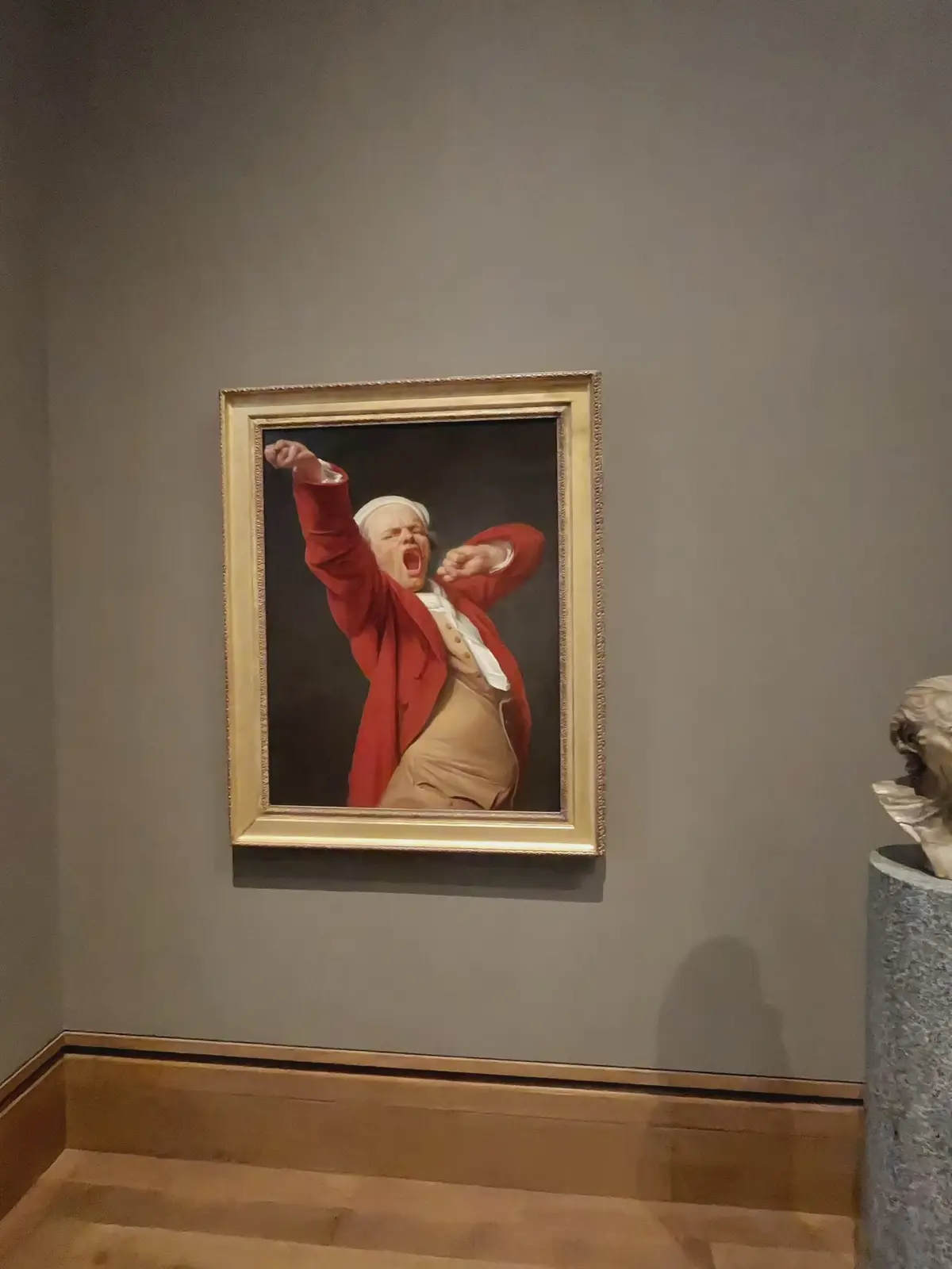 A classical painting of a man in a red coat and white wig, framed in gold, is hung on a gray museum wall. The man is…