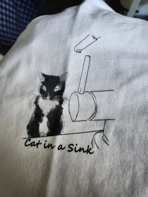 A close-up of a light-colored t-shirt featuring a black-and-white illustration of a cat sitting beside a sink with a faucet…