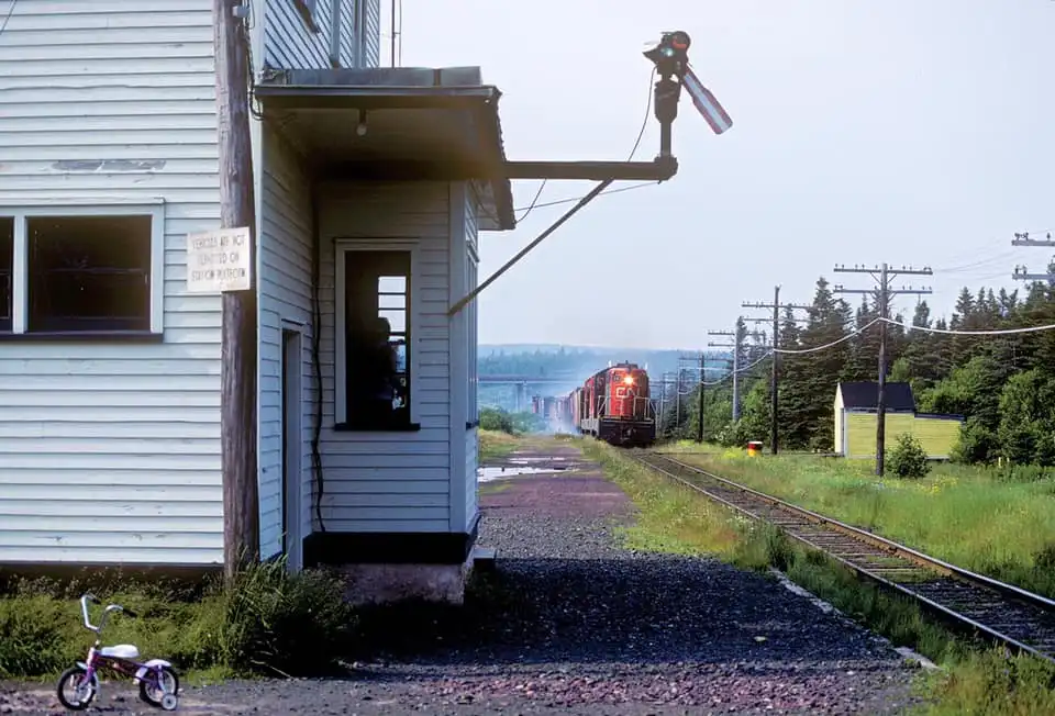 A vintage photo shows a small, weathered train station with a red train approaching on the tracks. A tiny pink tricycle sits…