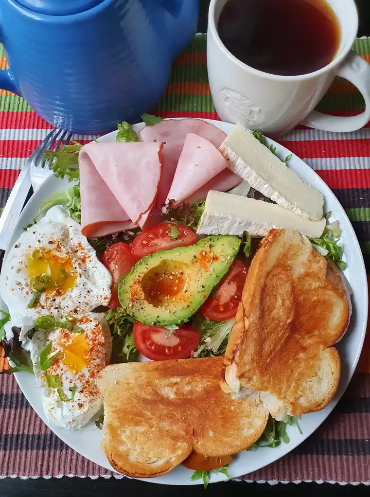 A white plate holds a colorful breakfast salad with poached eggs, avocado, tomato slices, ham, cheese, and toasted bread…